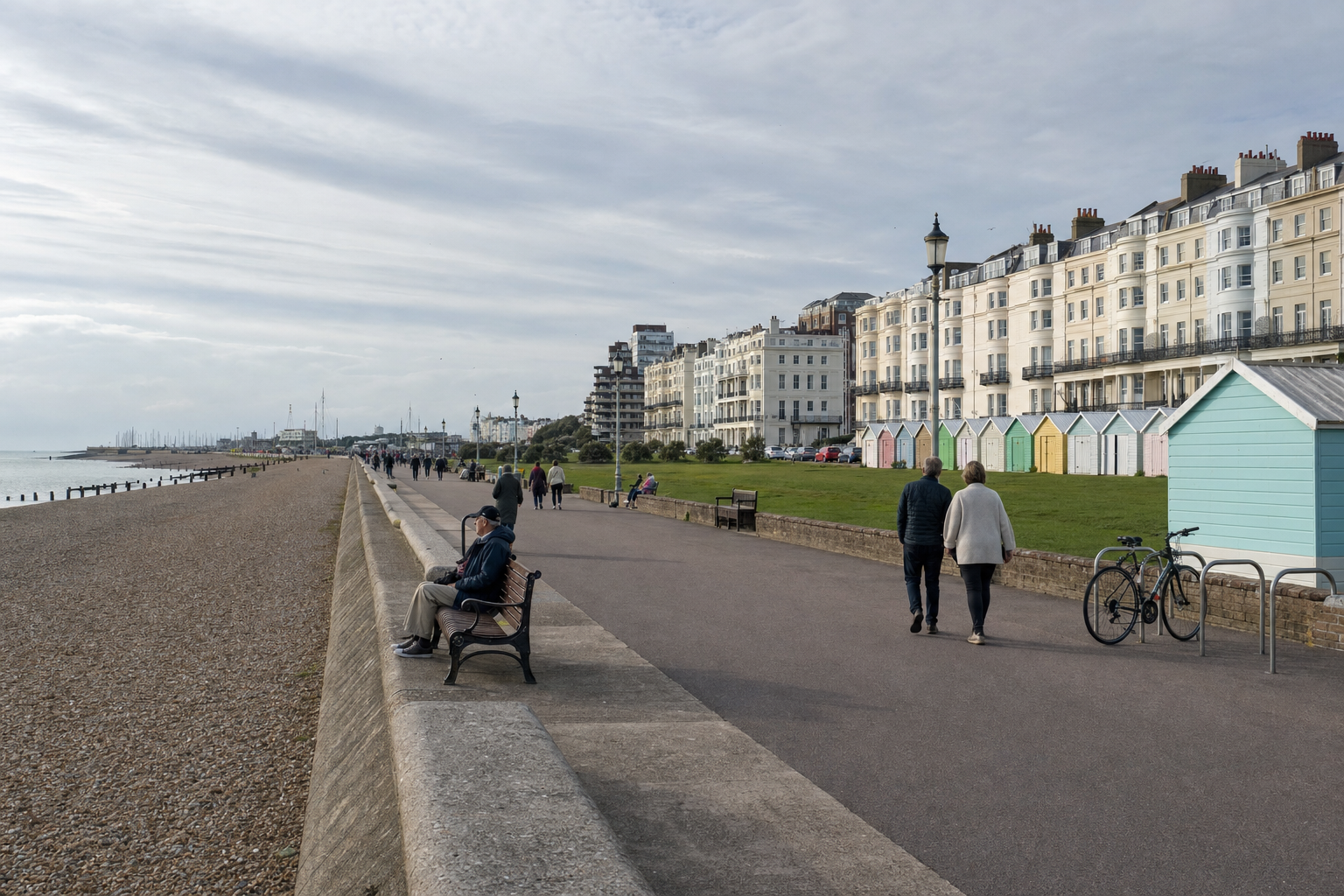People walking along a seaside promenade with colourful beach huts and Victorian-style buildings in the background.