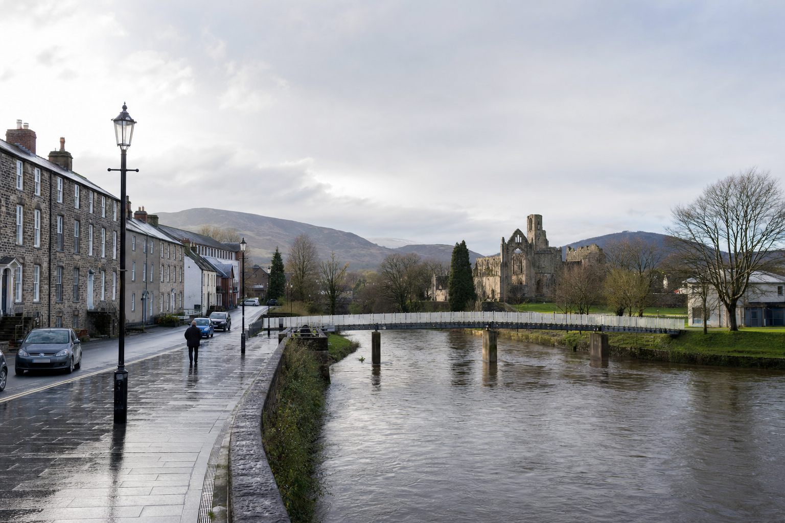 A solitary figure walks along a wet street beside a river with a picturesque bridge and historic ruins in the background.