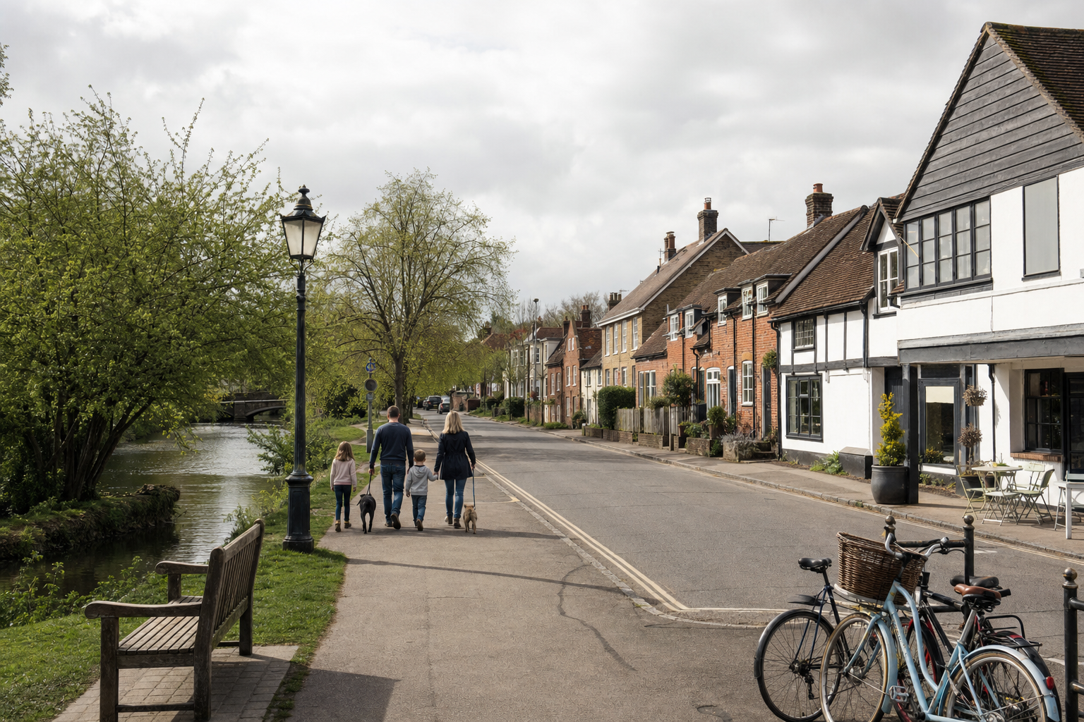Family walking with dogs along a riverside path beside a row of quaint houses and parked bicycles.