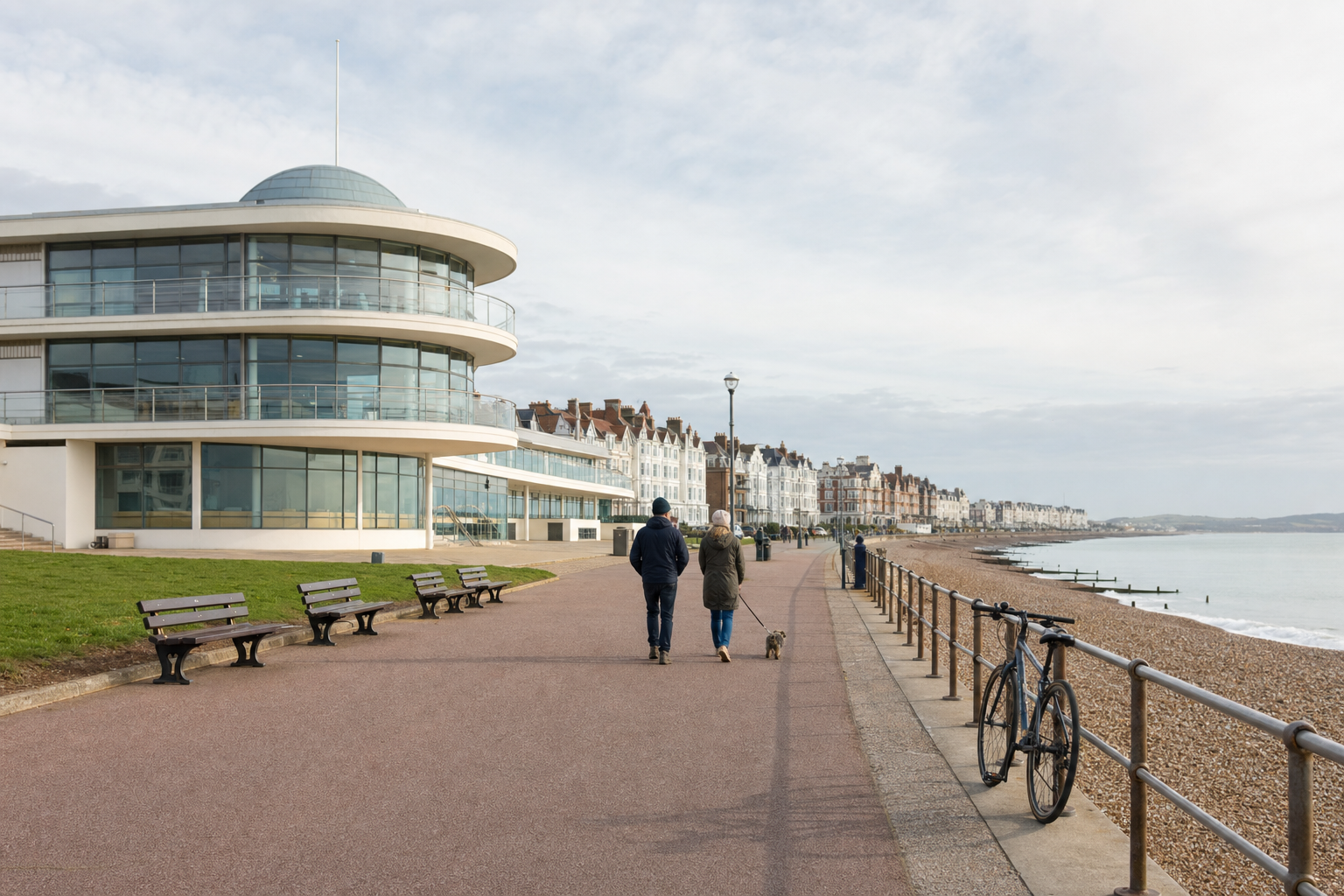 A couple walks a dog along a coastal promenade near a modern building with sea views.