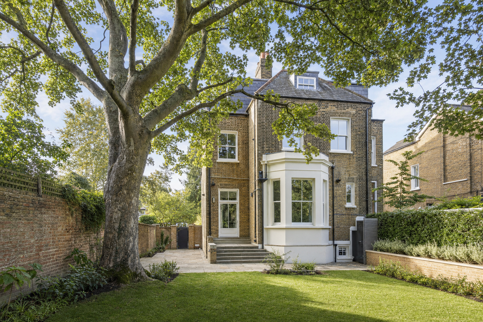 A large tree shades a Victorian-style brick house with a manicured garden and pathway.