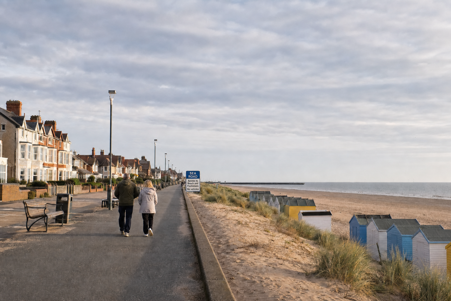 A couple strolls along a seaside promenade with colourful beach huts and residential houses in the background.