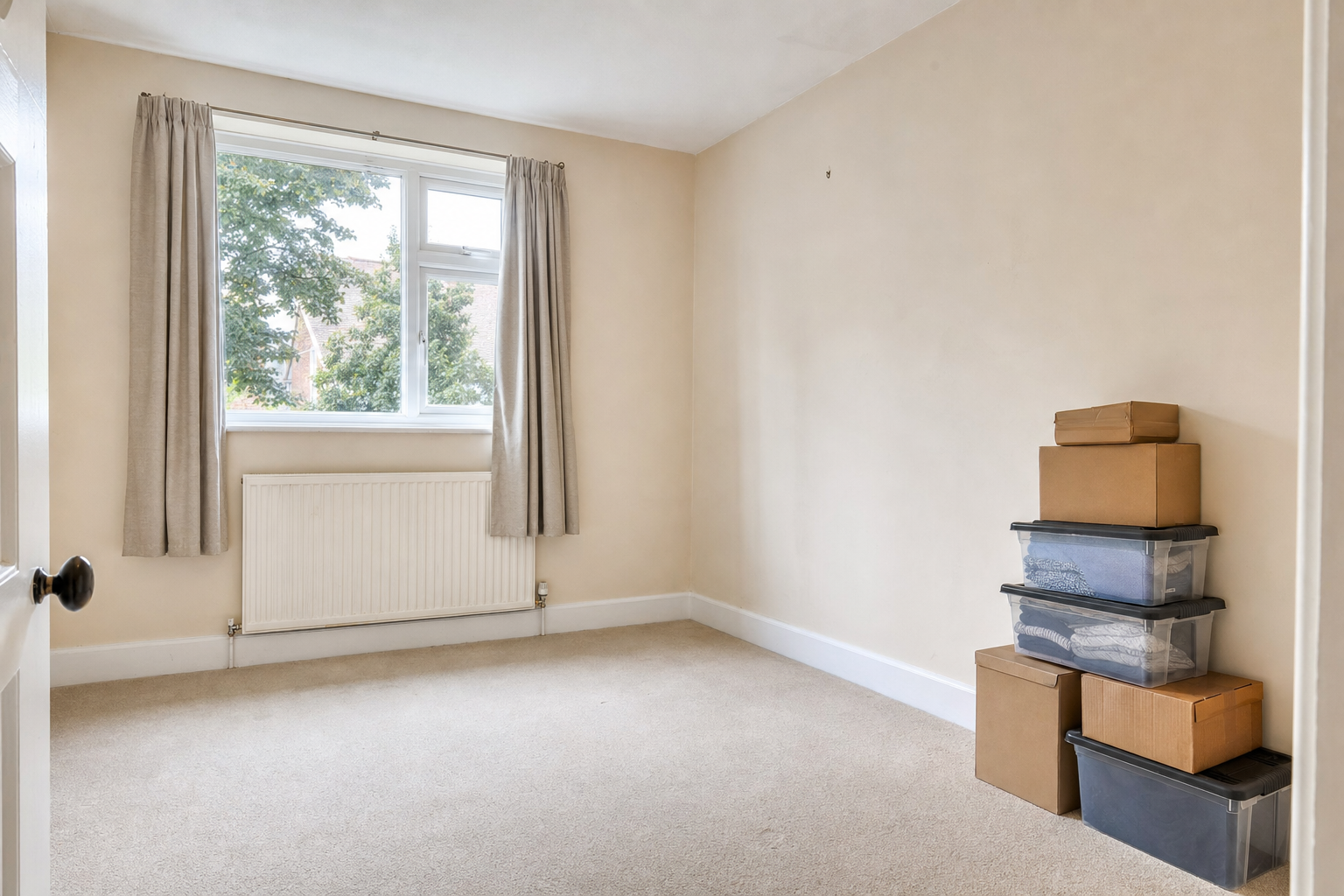 Unfurnished beige room with a window, curtain, radiator, and stacked cardboard and plastic boxes.