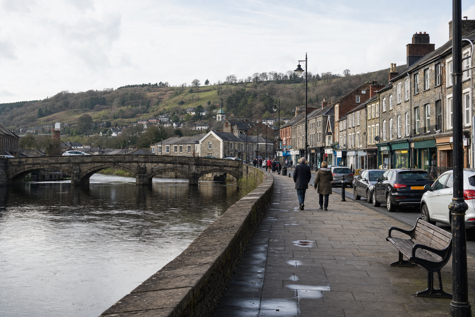 A couple walks along a riverside pavement in a quaint town, with a stone bridge and hills in the background.