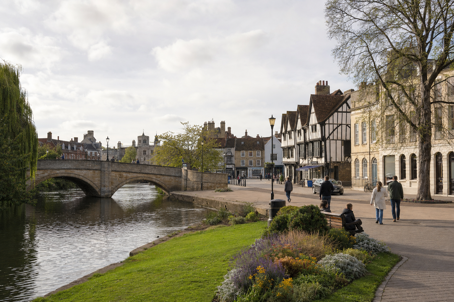 A scenic riverside scene in a historic town with a stone bridge and people strolling on a sunny day.