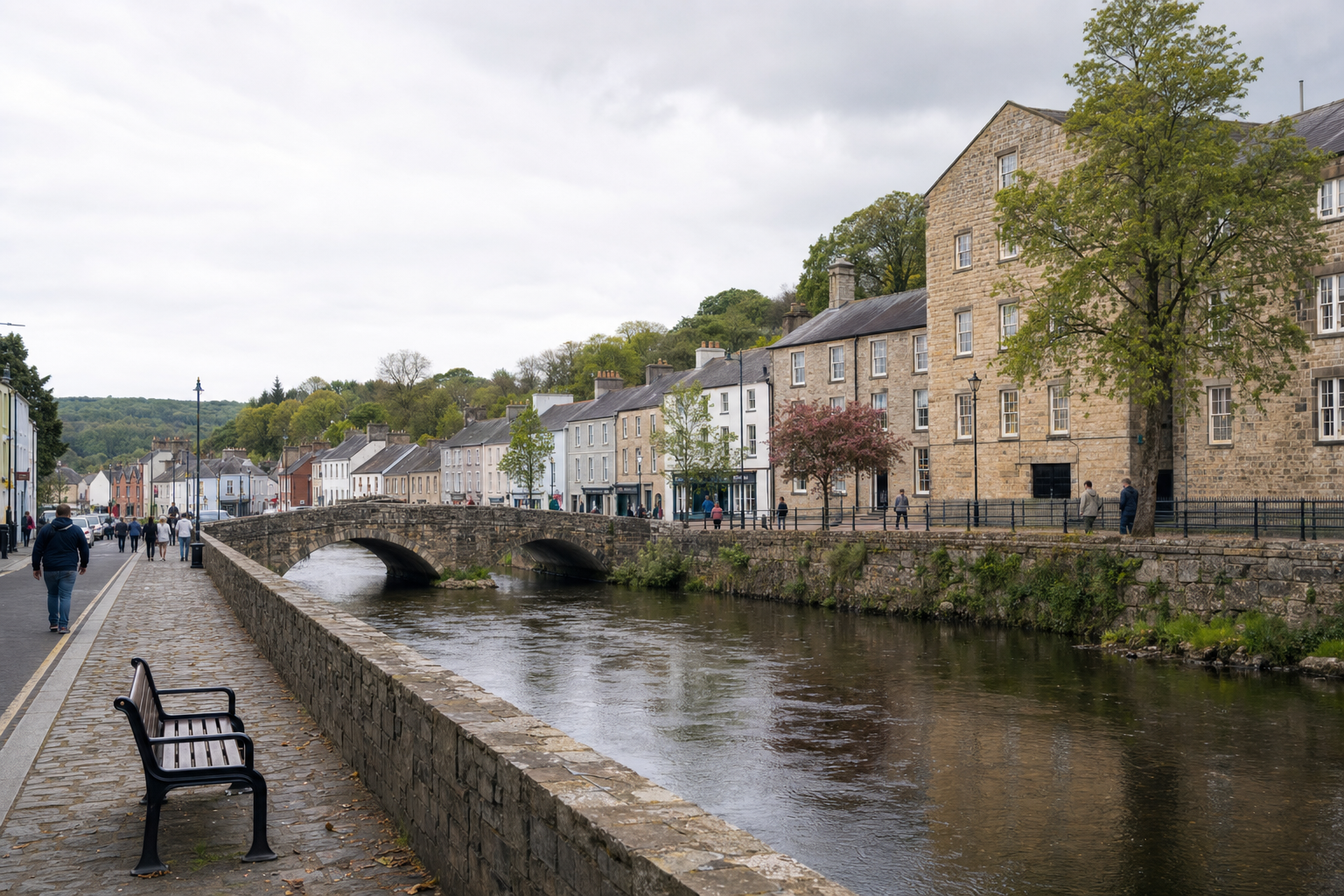 A quaint riverside town with historic stone bridge, colourful buildings, and people strolling along the path on a cloudy day.