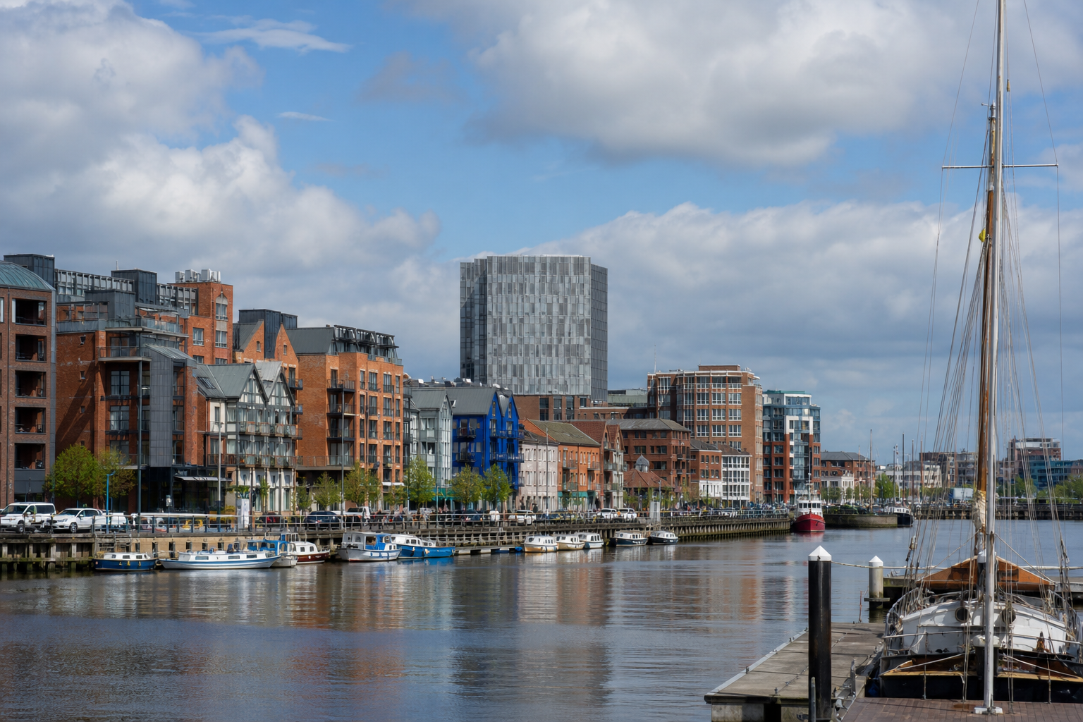 Riverside view of boats docked alongside modern brick buildings under a partly cloudy sky.