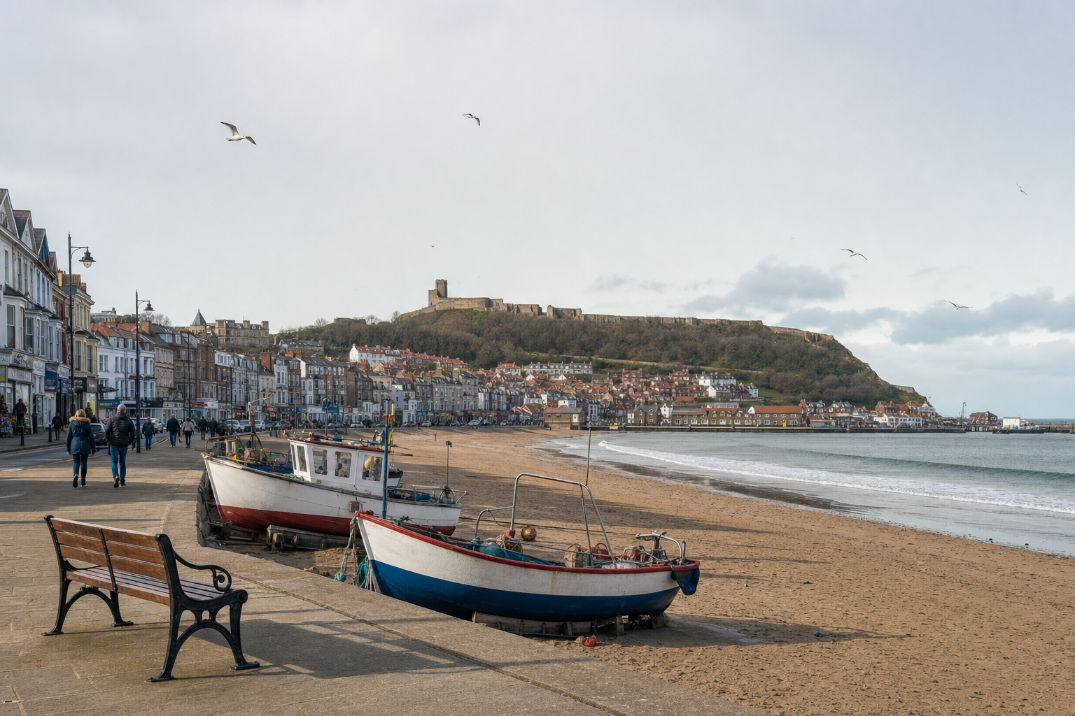 Seaside town with boats on the beach, a castle on the hill, and people walking along the promenade.