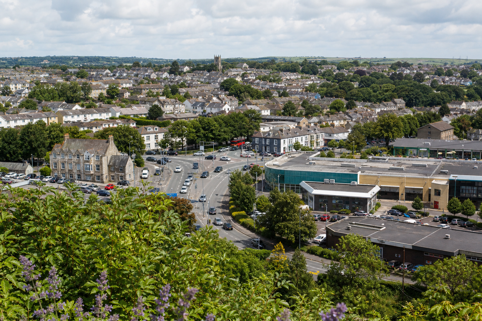 Aerial view of a suburban town with roads, parked cars, and greenery under a partly cloudy sky.
