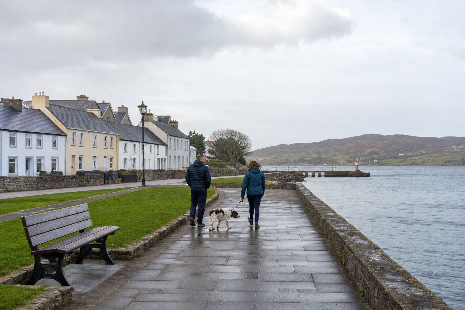 Two people walking a dog along a waterfront pathway in a quaint village.