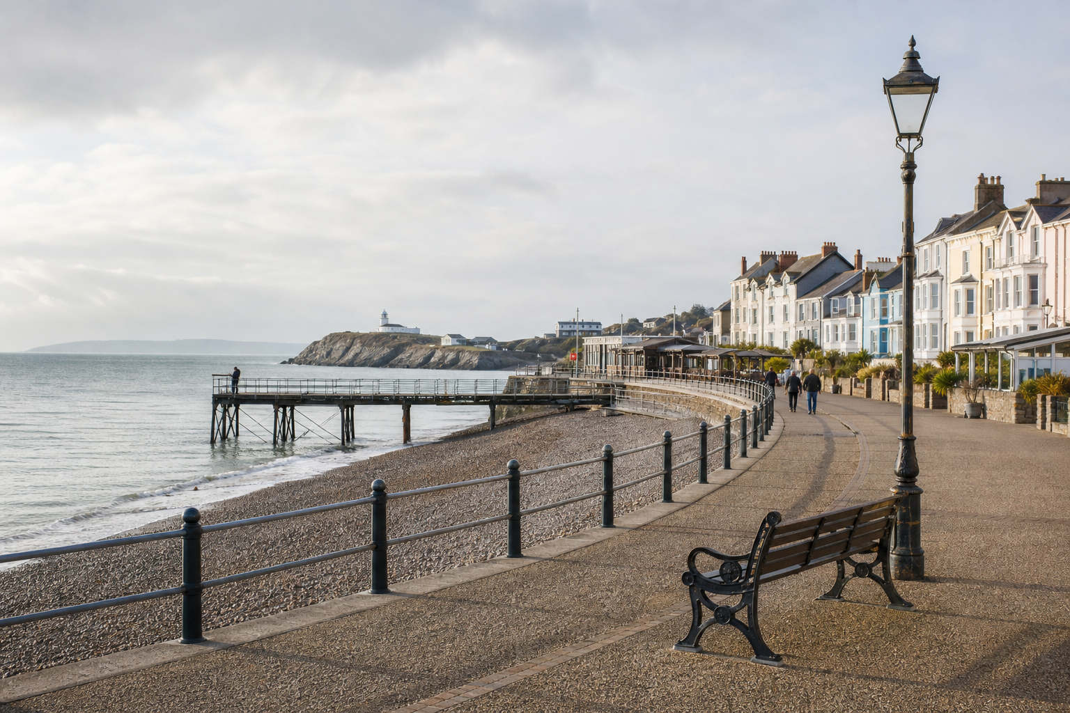 Seaside promenade with a bench, pebbled beach, pier, and rows of traditional houses under a cloudy sky.