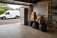 A garage with packed boxes, a bicycle, and bin bags, while a white van is parked outside on the driveway.