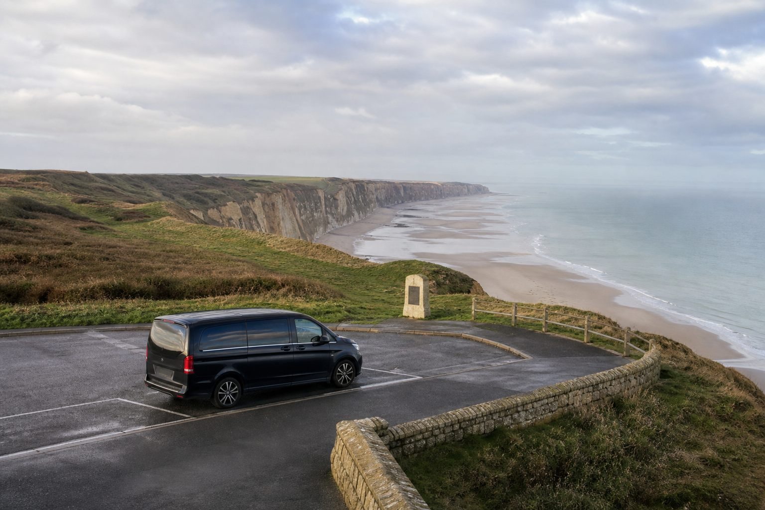 A black van parked at a coastal viewpoint overlooking a sandy beach and cliffs under a cloudy sky.