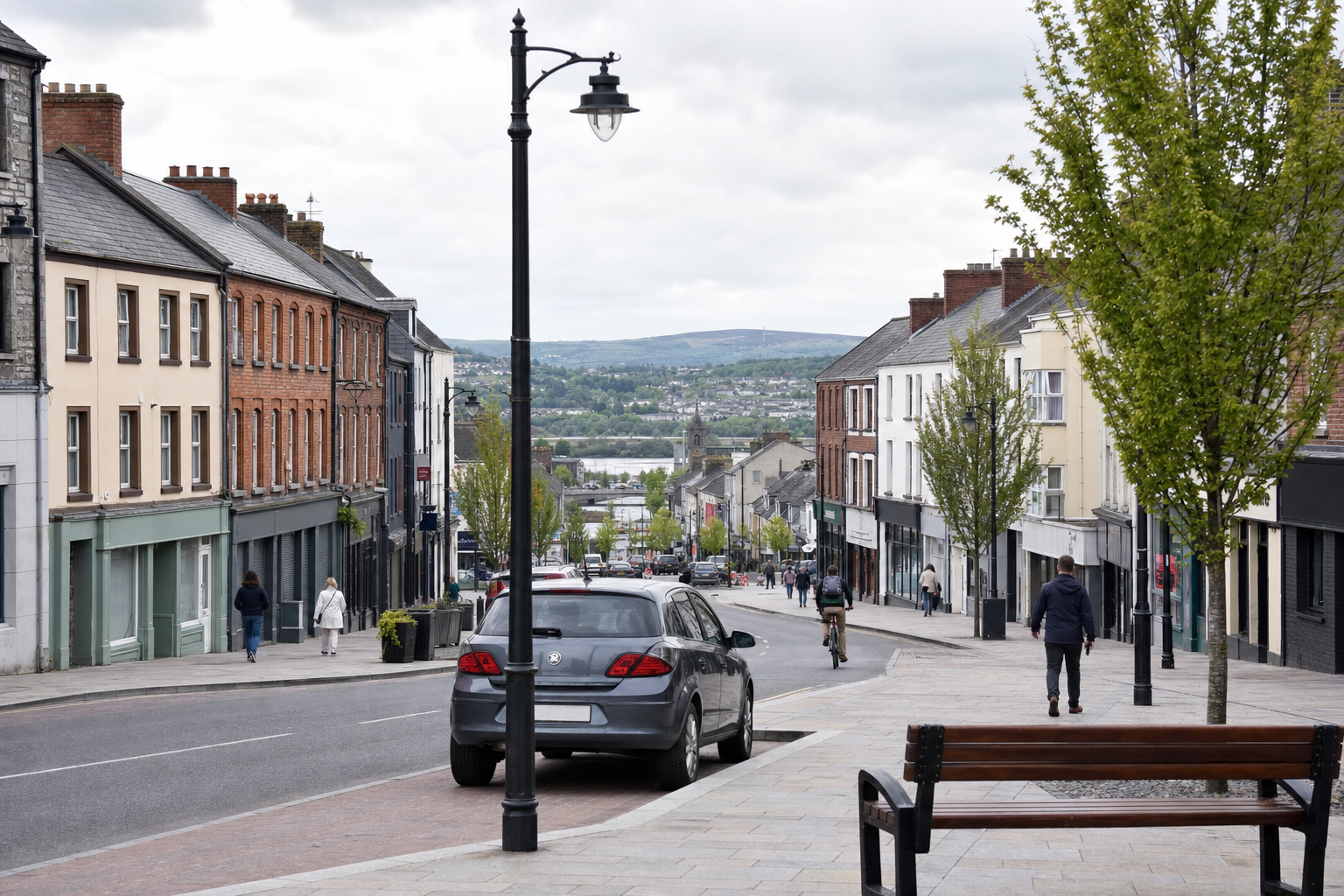 A sloping street lined with buildings, trees, and a view of a distant river, with people walking and a parked car.