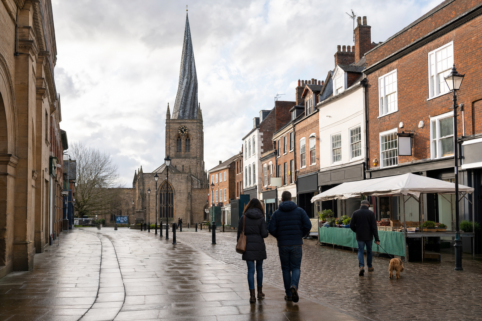 People walk along a wet, cobbled street beside shops, approaching a church with a tall spire, under a cloudy sky.