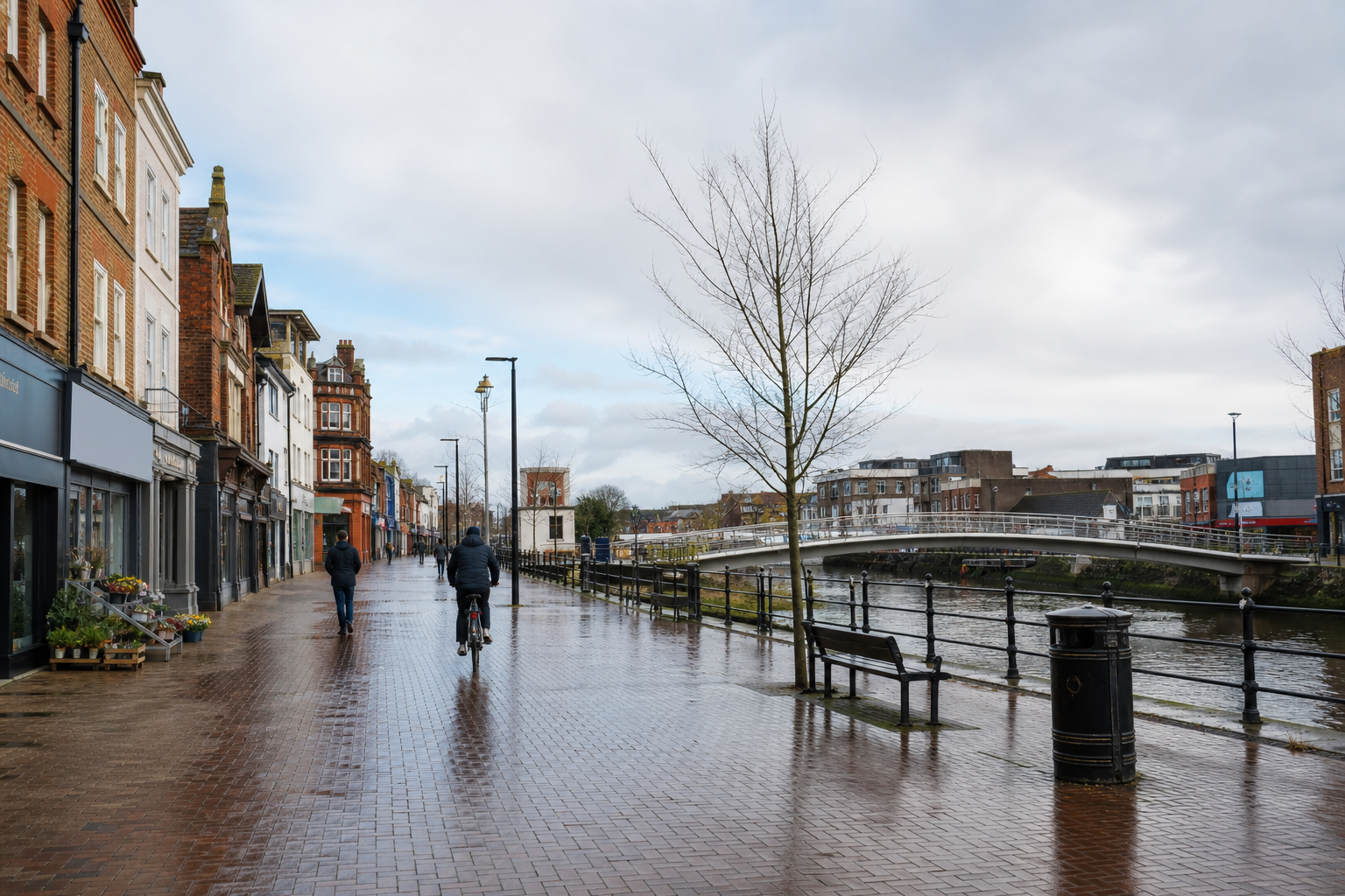 A riverside street lined with shops and trees, featuring a cyclist and pedestrian under a cloudy sky near a bridge.