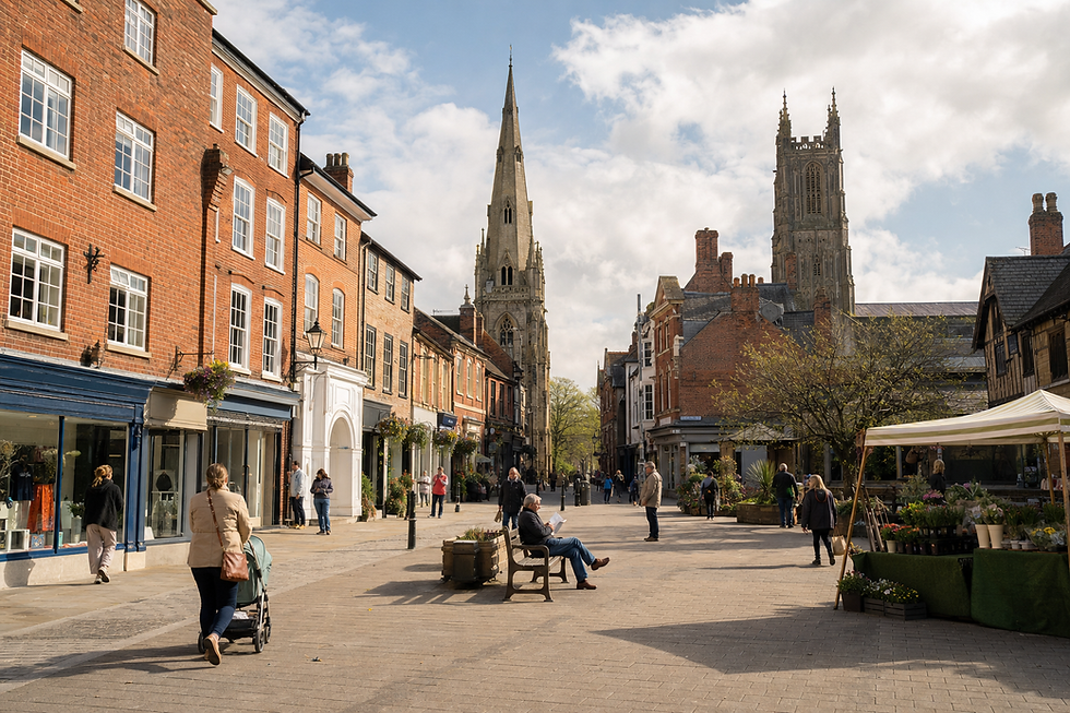 People strolling along a charming high street with historic buildings and a prominent church spire in the background.