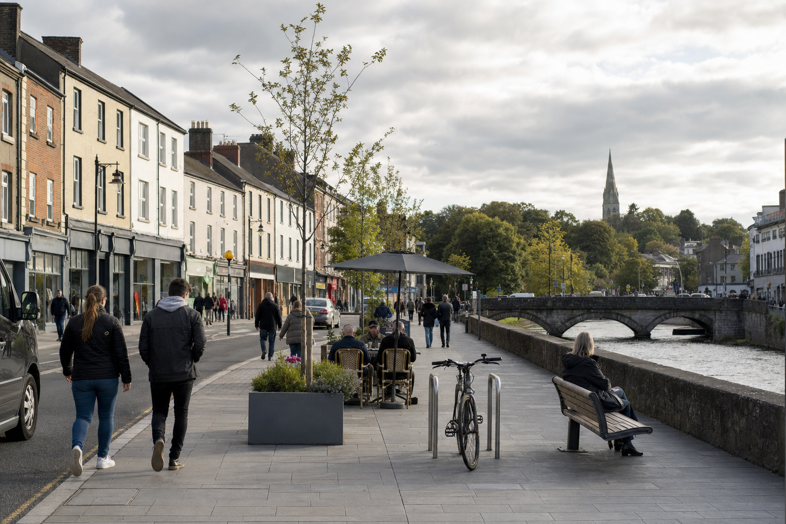 People strolling along a riverside promenade with outdoor seating, buildings, and a stone bridge in the background.