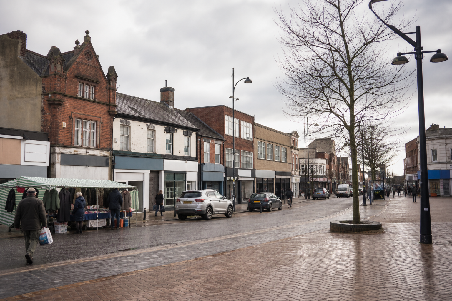 A grey, overcast day on a British high street with a market stall and pedestrians strolling along the wet pavement.