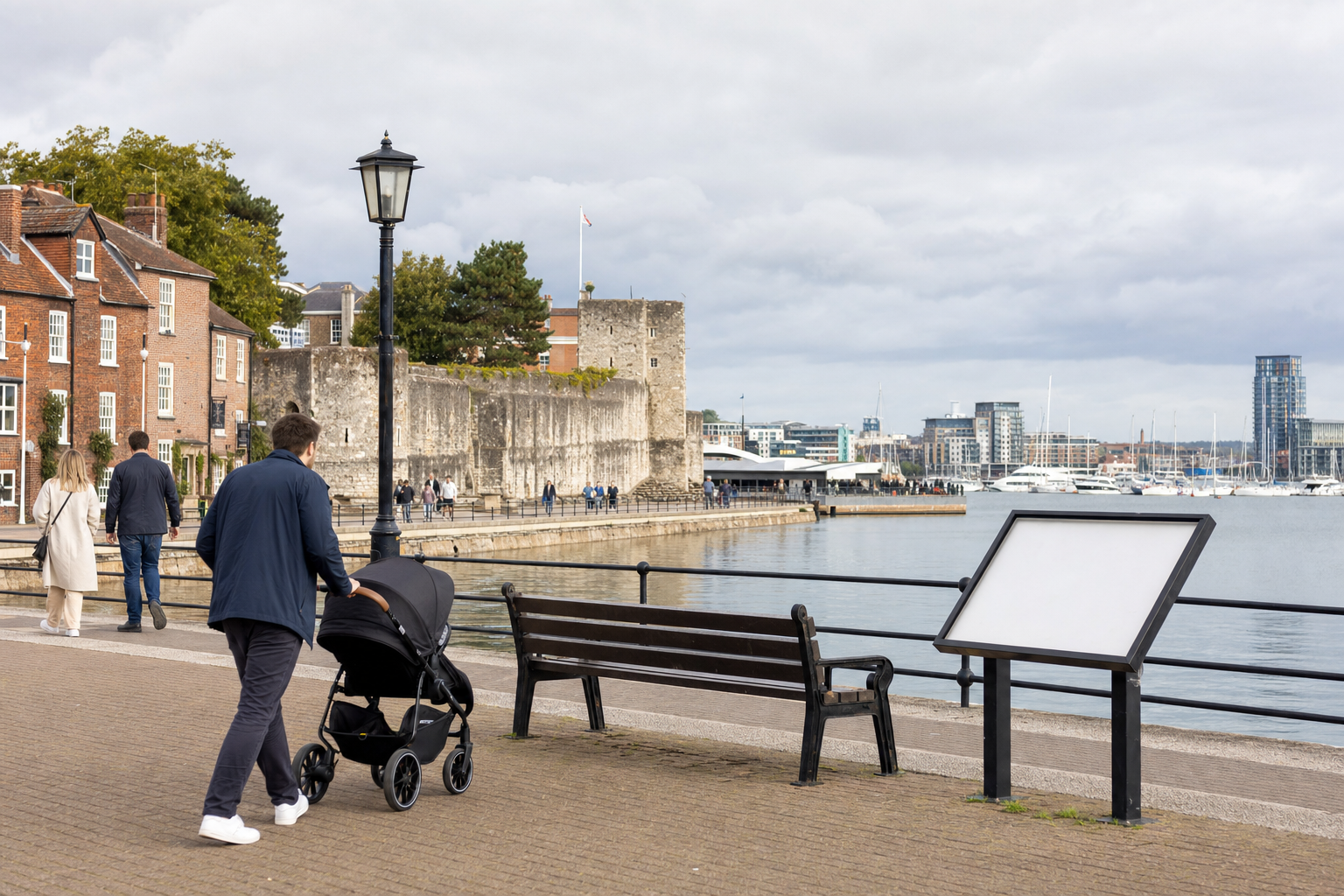 A person pushes a pram along a waterfront promenade with historic buildings and docked boats in the background.