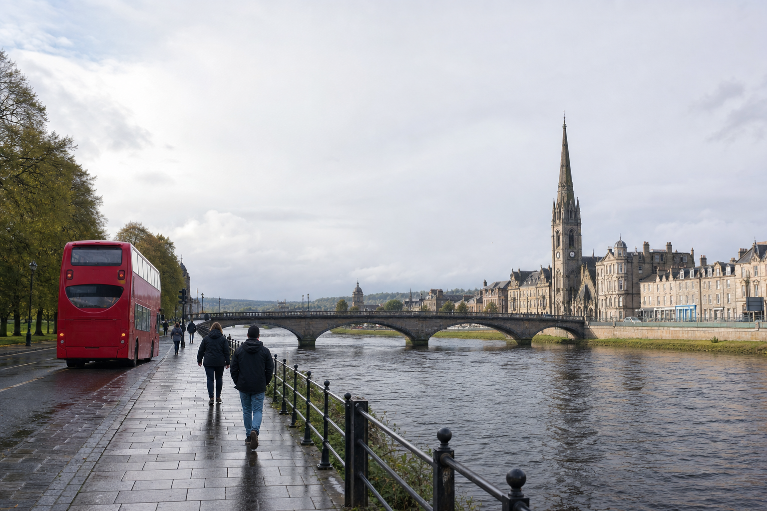 A red double-decker bus drives along a riverside road lined with trees, facing a historic town and bridge under a grey sky.
