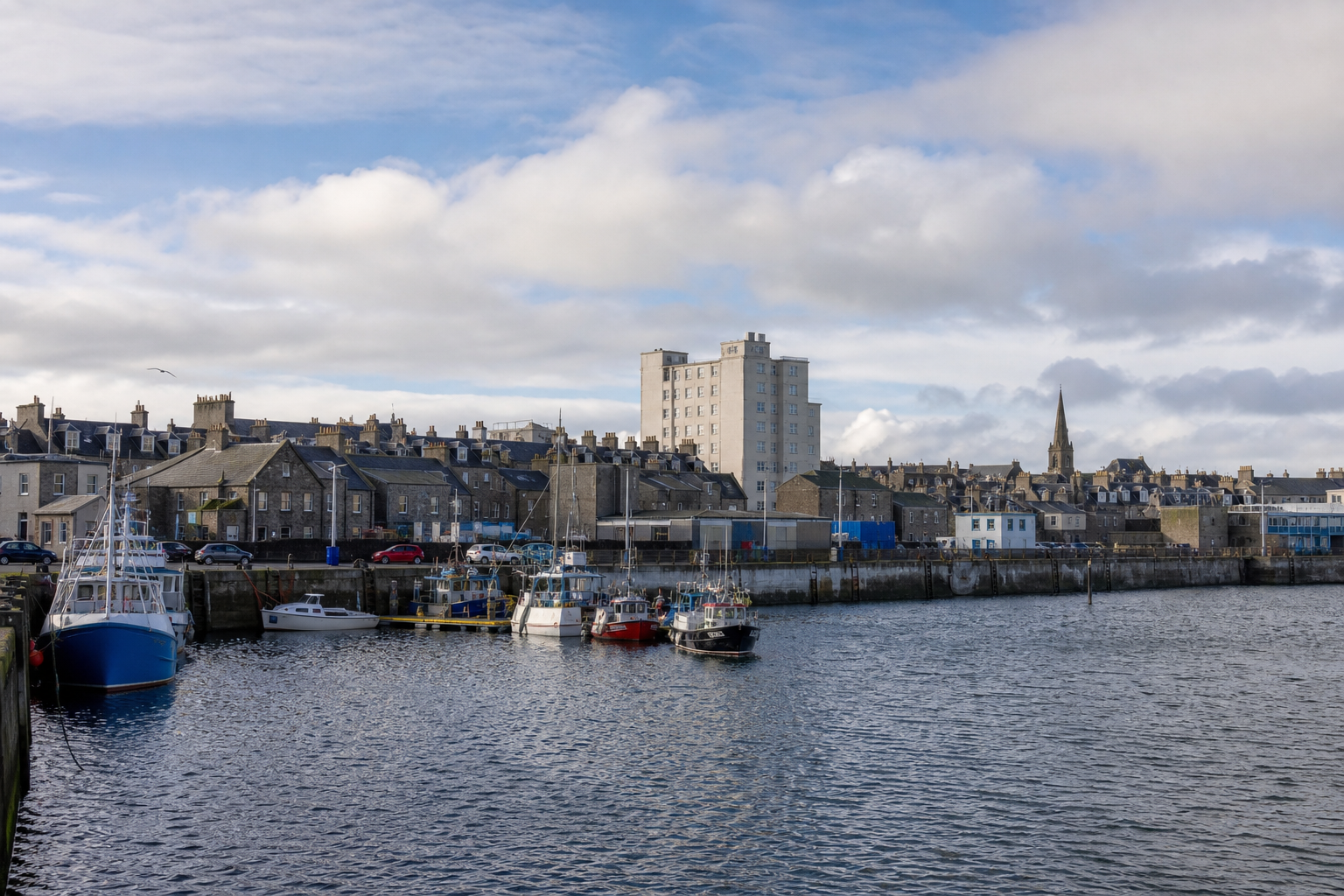Harbor with fishing boats, historic buildings, and a tall apartment block under a partly cloudy sky.