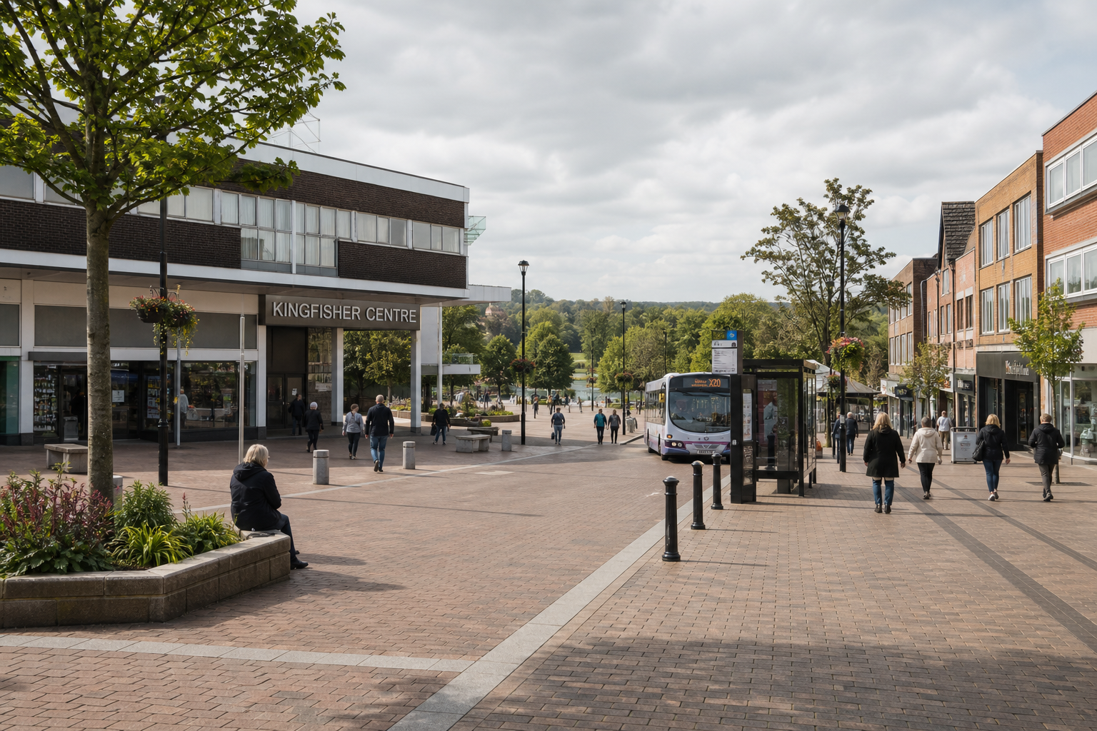 Pedestrians walk near the Kingfisher Centre in a shopping area with a bus stop and surrounding greenery.