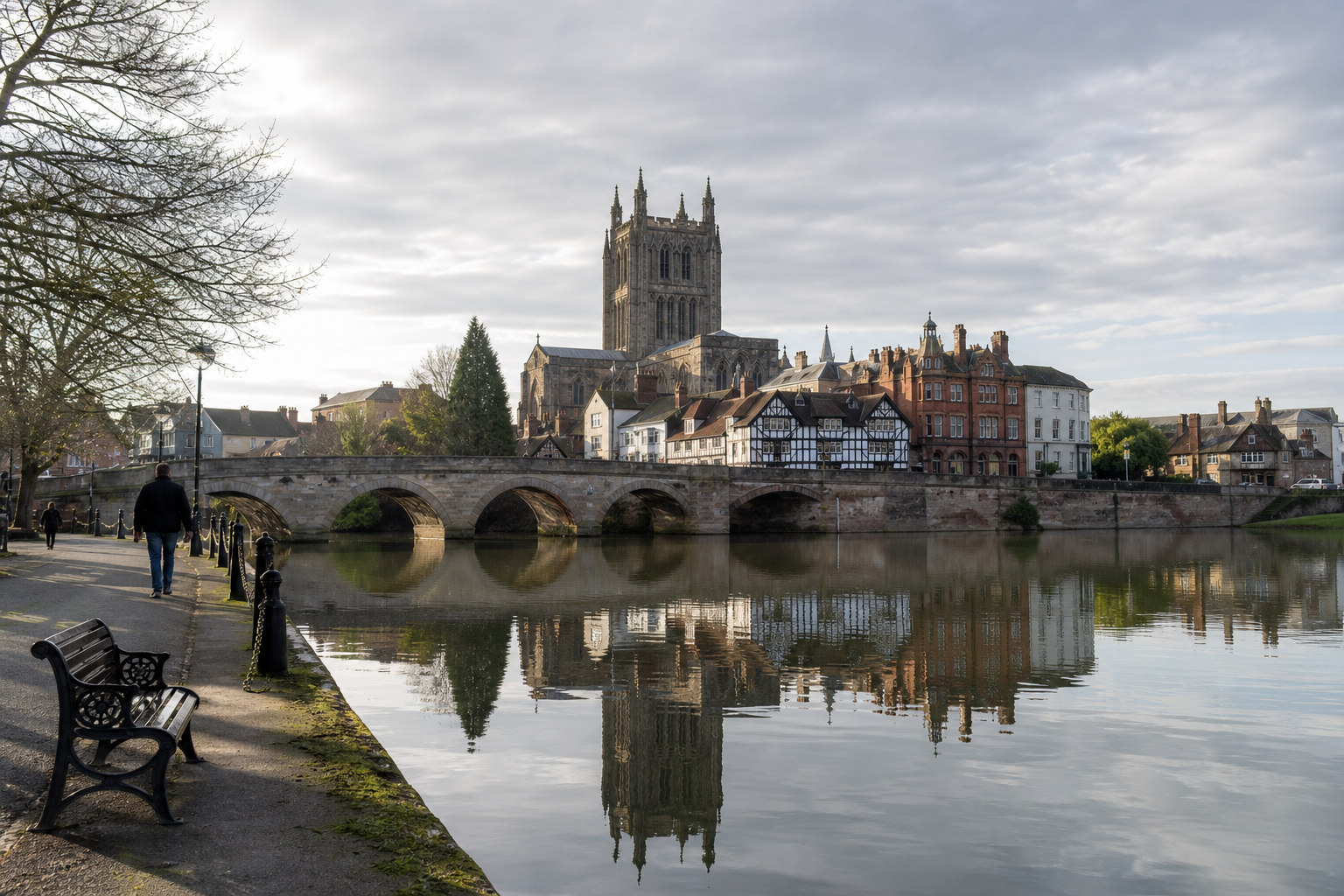 Historic bridge and cathedral reflected in a calm river, with a tree-lined pathway and benches in the foreground.