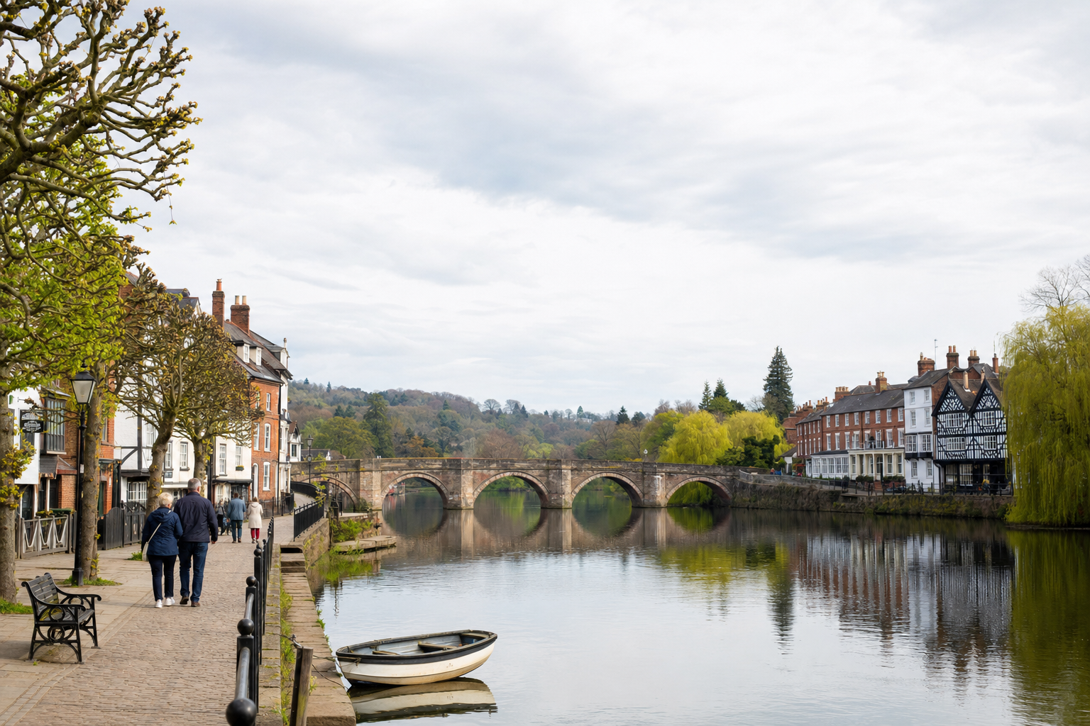 Riverside path with people strolling alongside historic buildings and a stone bridge reflecting in the water.