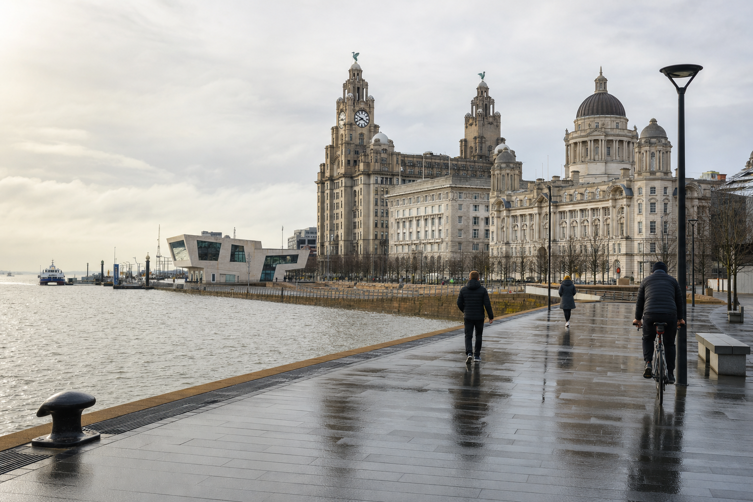 People walking and cycling along Liverpool's waterfront with iconic historical buildings in the background on a cloudy day.