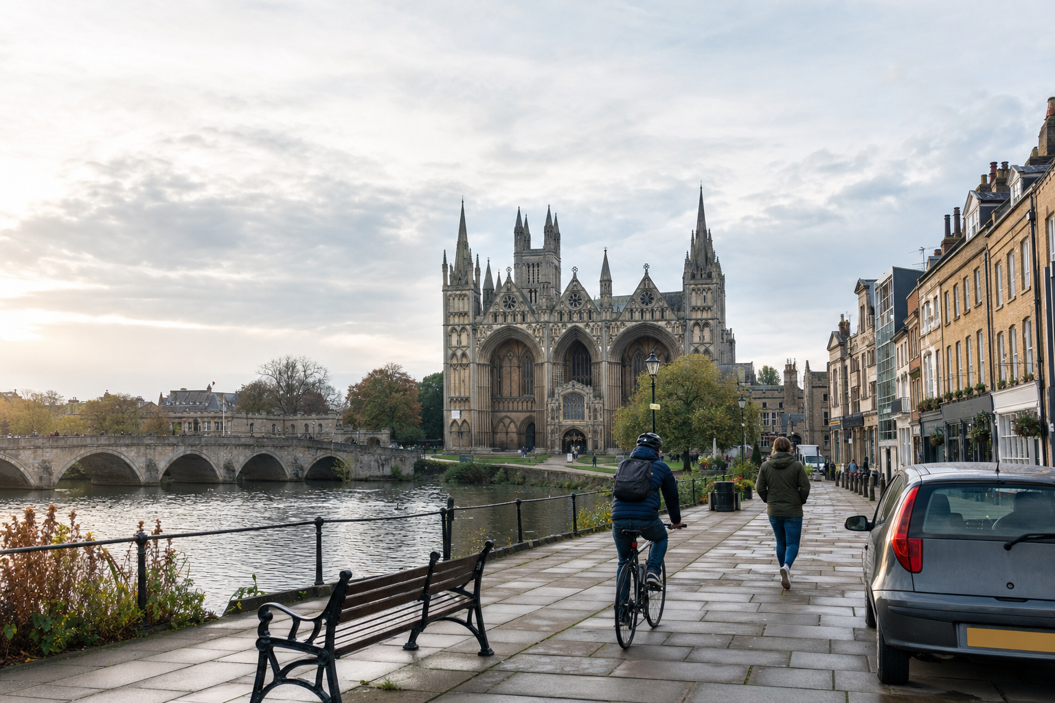 A cyclist and pedestrian on a riverside path near a grand Gothic cathedral with an arched bridge in the background.