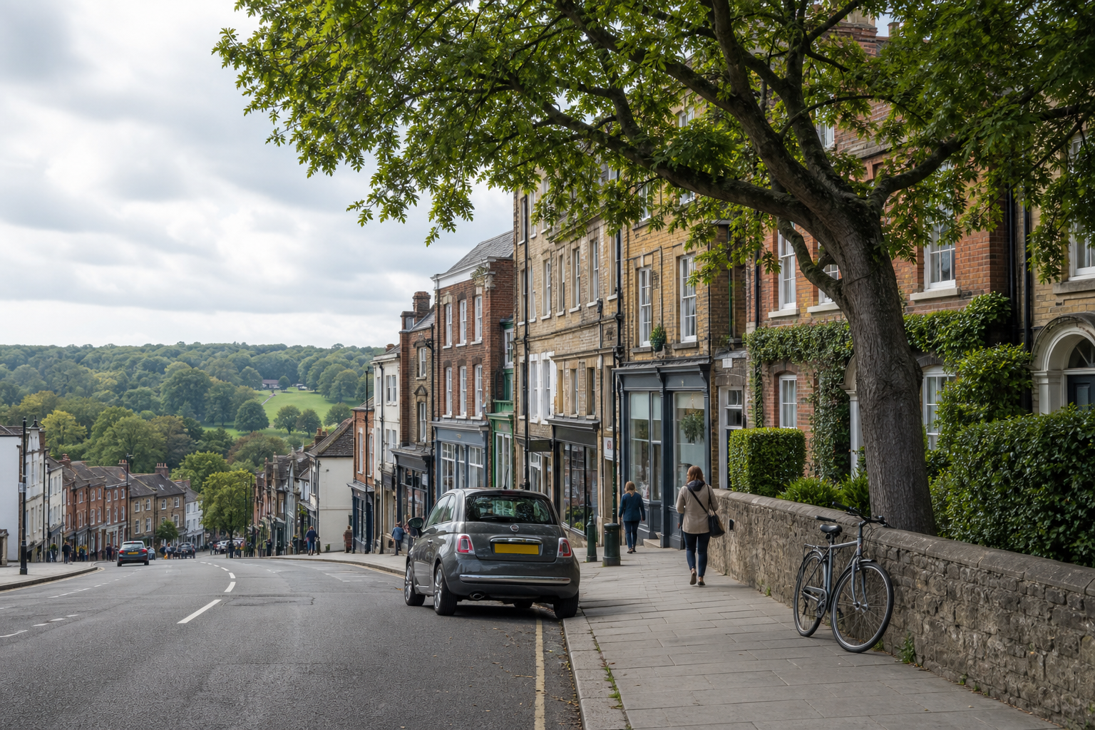 A sloping street lined with historic brick buildings, a parked car, pedestrians, and a bicycle, with green fields in the background.