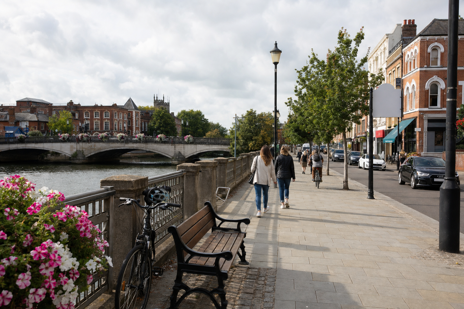 People walking along a riverside path, with a bicycle parked by a bench and a historic townscape in the background.