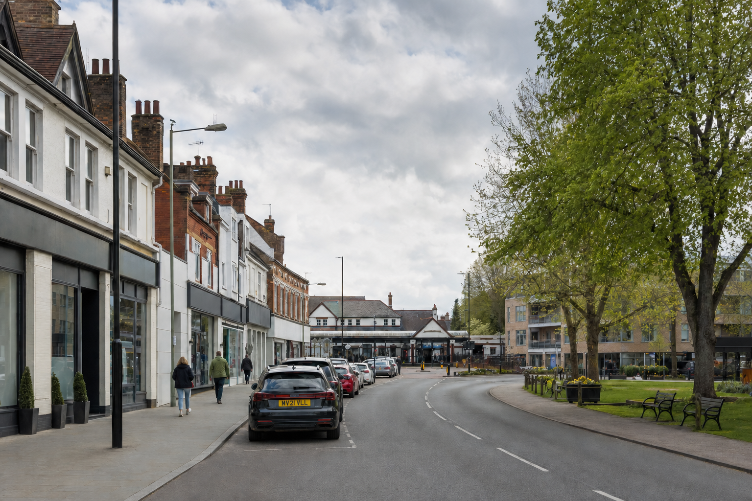 A curved street lined with shops and parked cars, bordered by a grassy area with benches and trees under a cloudy sky.