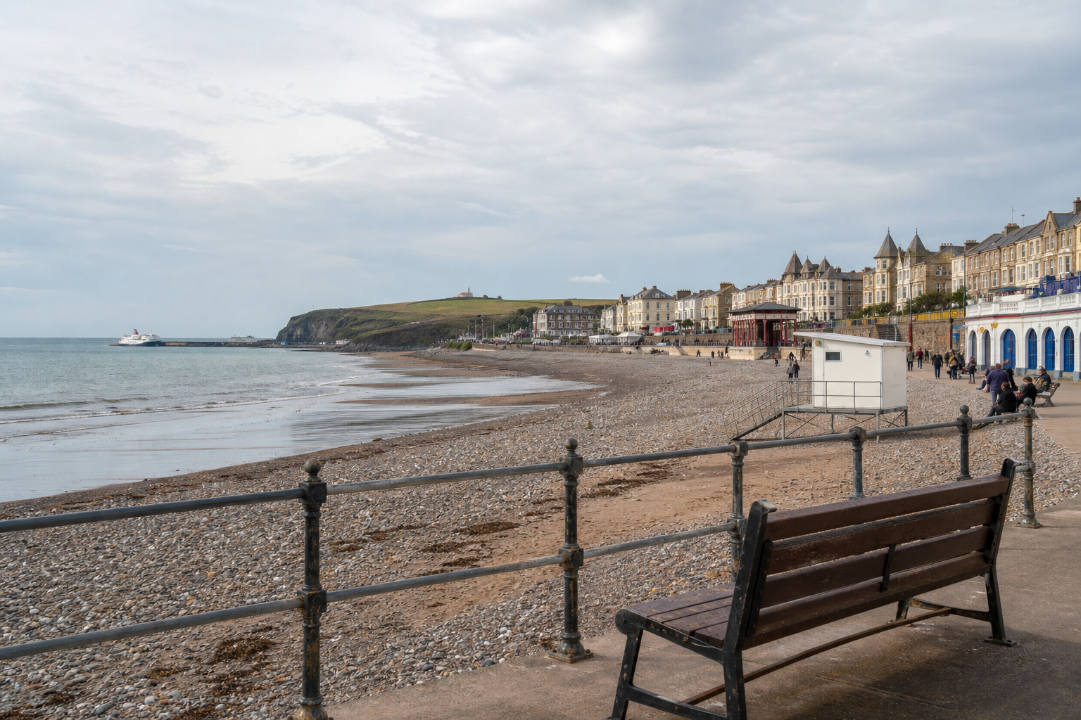 View of a pebble beach and promenade with Victorian buildings, railing, bench, and ship in the distance under a cloudy sky.