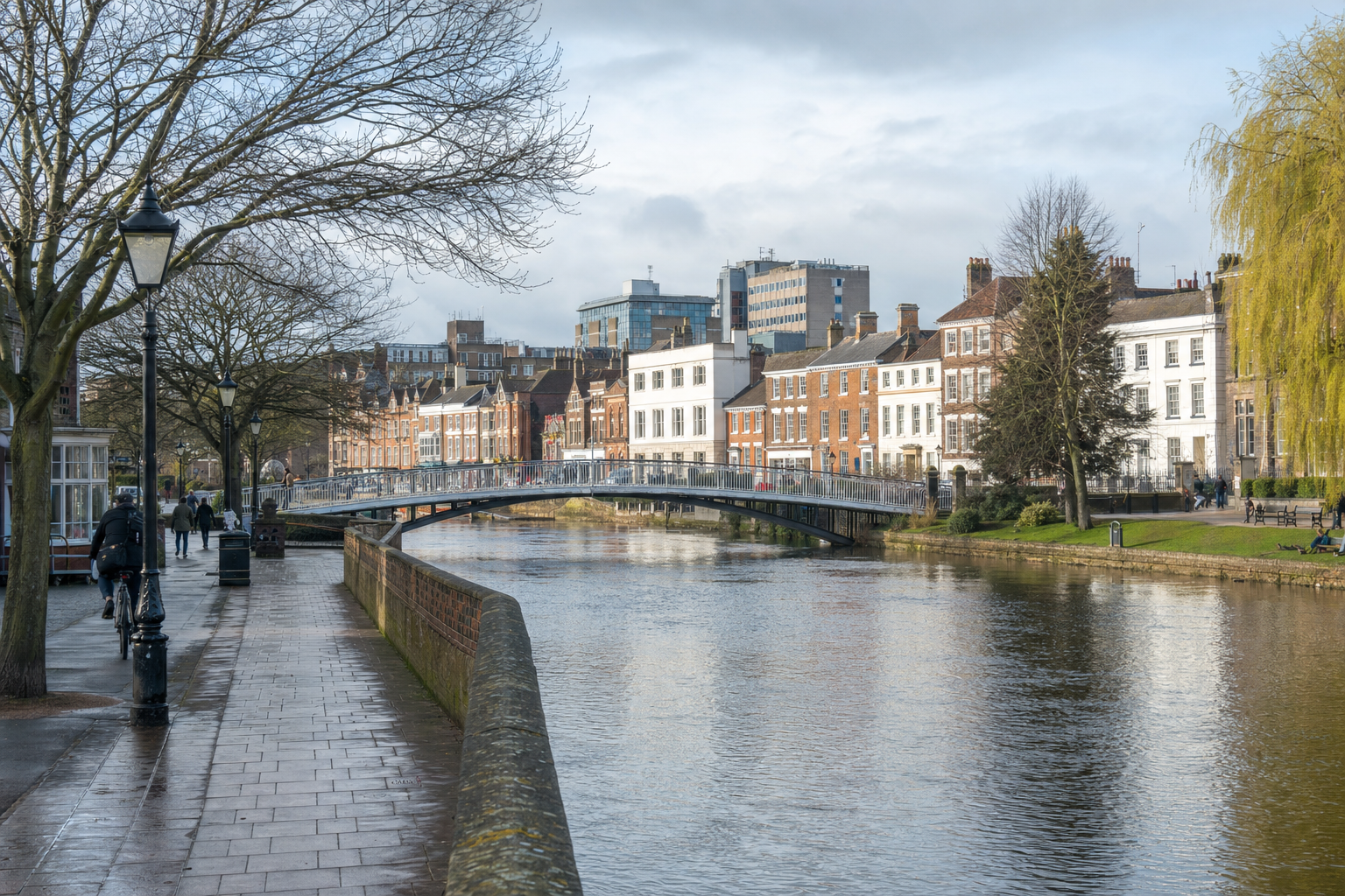 A tranquil canal lined with historic buildings, a pedestrian bridge, and people walking or cycling along the path.