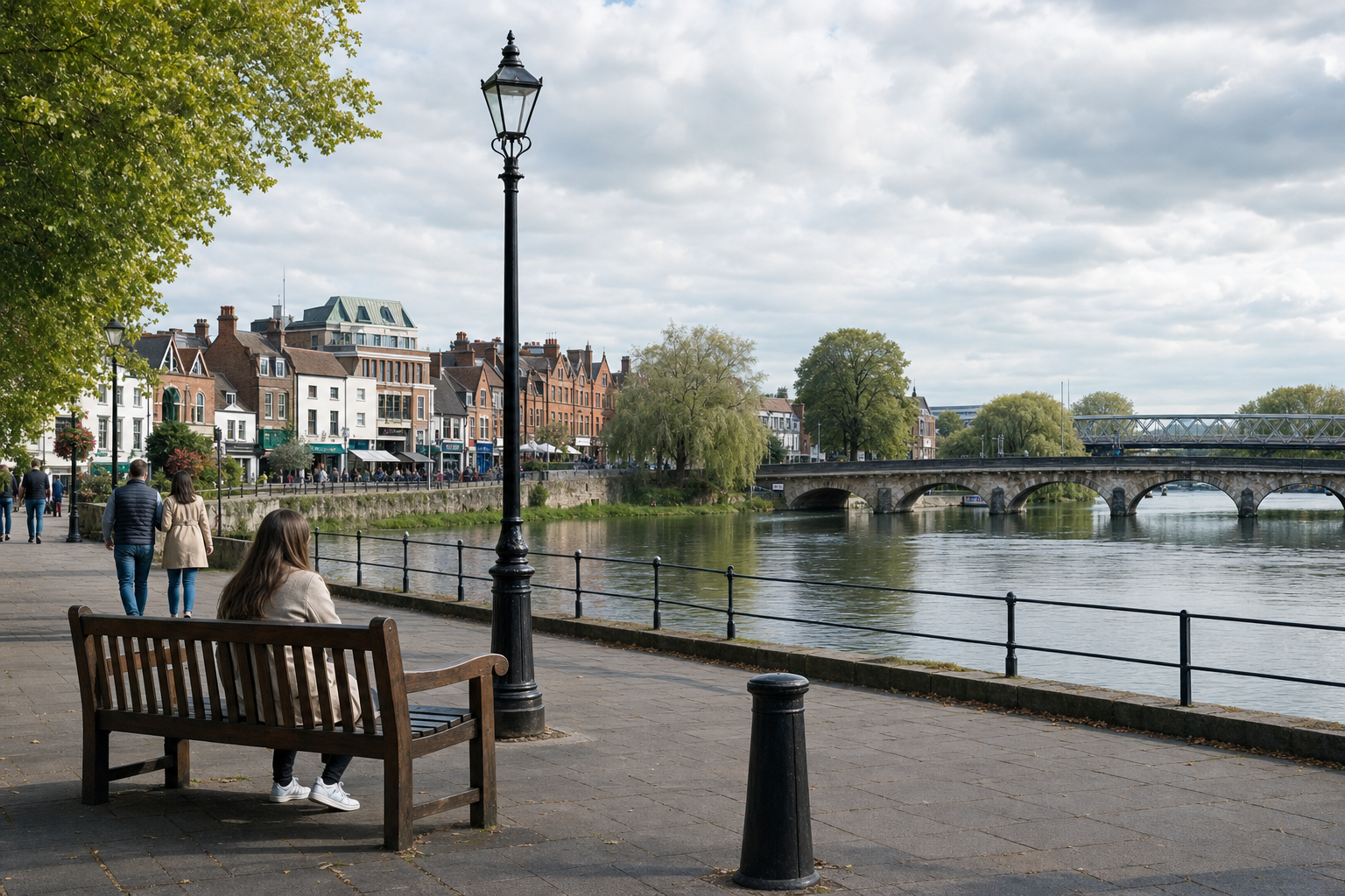 A woman sits on a bench overlooking a scenic riverside path with historic buildings and a bridge in the background.