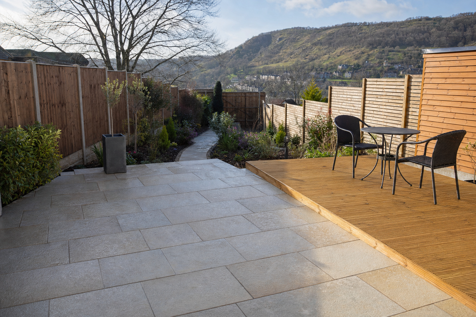 Contemporary garden patio with paving, a wooden deck, and a table with two chairs, overlooking a hilly landscape.