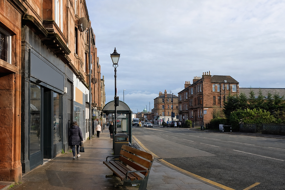 A quiet street scene features red sandstone buildings, a bus shelter, and pedestrians walking along the pavement.