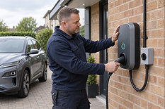 A man in workwear installs a charging station outside a house next to a parked electric vehicle.