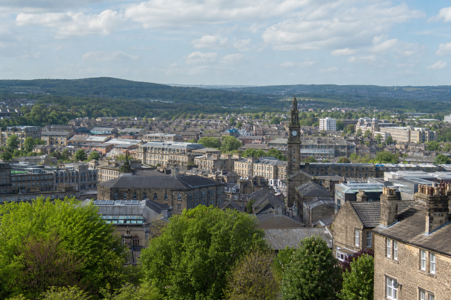 Aerial view of a town with a prominent clock tower, surrounded by green hills under a partly cloudy sky.