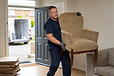 A man in gloves carries an armchair into a house from a van parked outside, with cardboard boxes nearby.