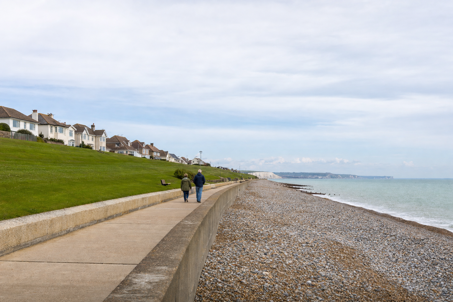 Two people walking along a coastal path with houses on a grassy hill and a pebble beach stretching alongside.