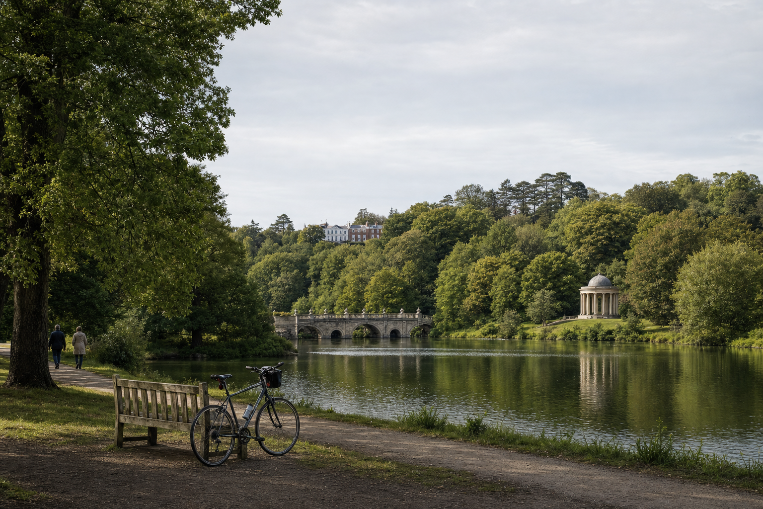 A bicycle leans against a bench by a lake, with a bridge and a small domed temple in the background amidst lush greenery.