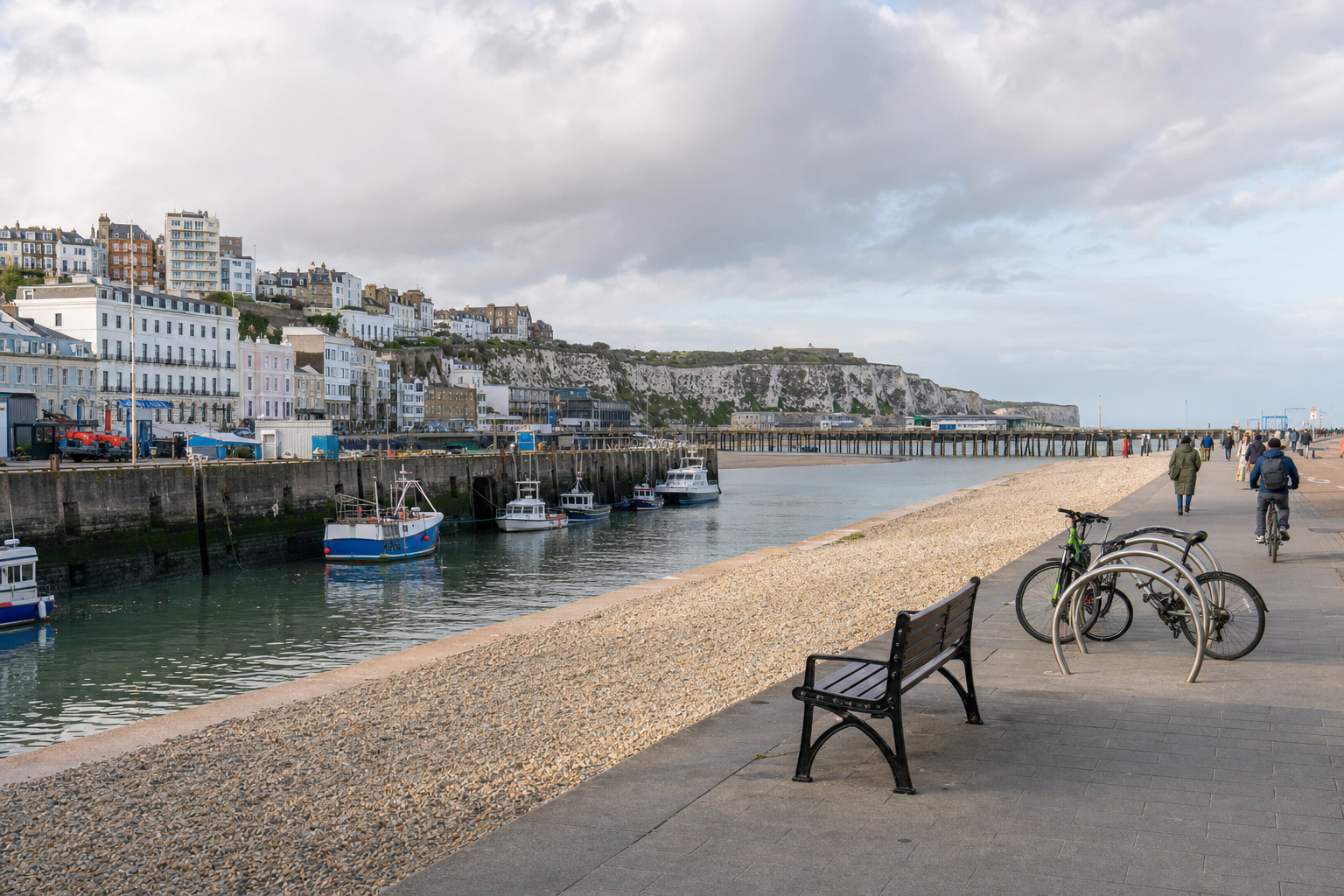 Fishing boats docked by the harbour, with people walking and cycling along a coastal promenade lined with white cliffs.
