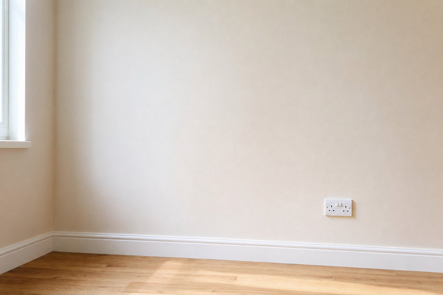 Sunlit corner of an empty room with wooden floor, featuring a wall socket and adjacent window.