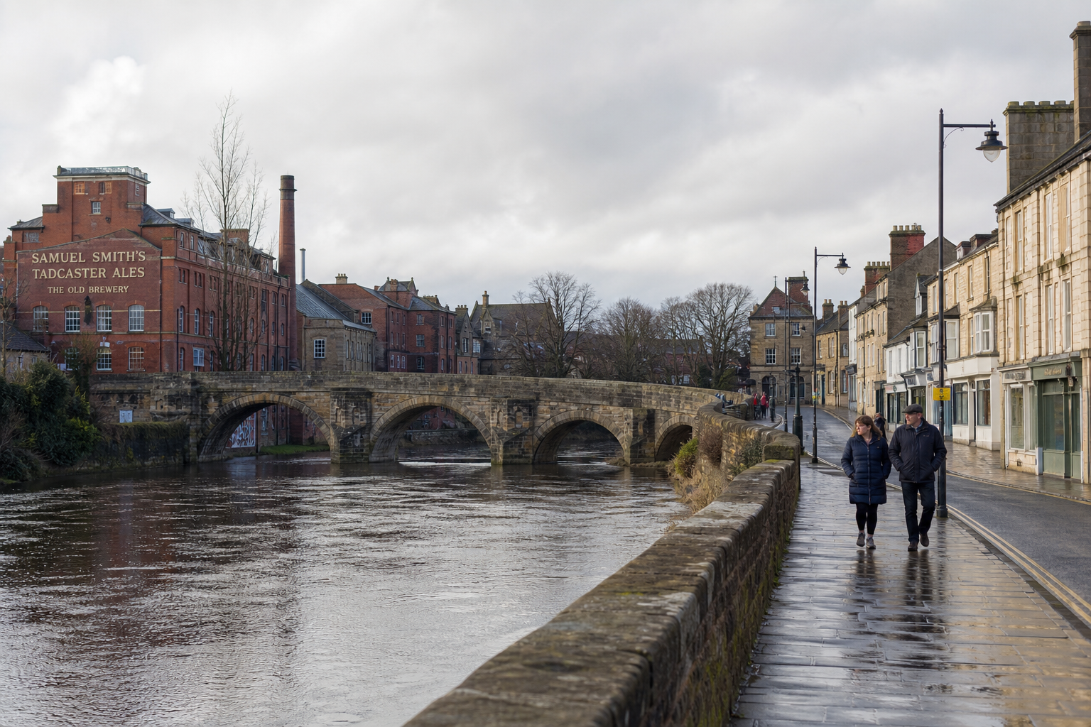 A couple walks along a riverside promenade in Tadcaster, with historic buildings and a stone bridge in the background.