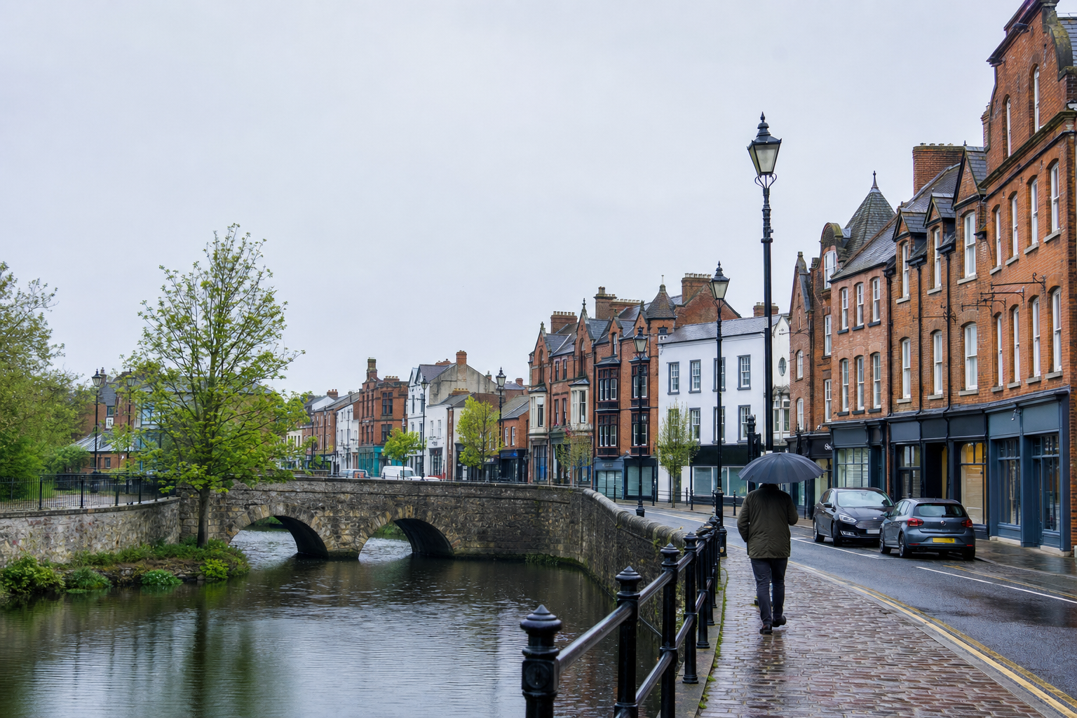 A person with an umbrella walks along a rainy riverside street lined with historic brick buildings and an arched stone bridge.
