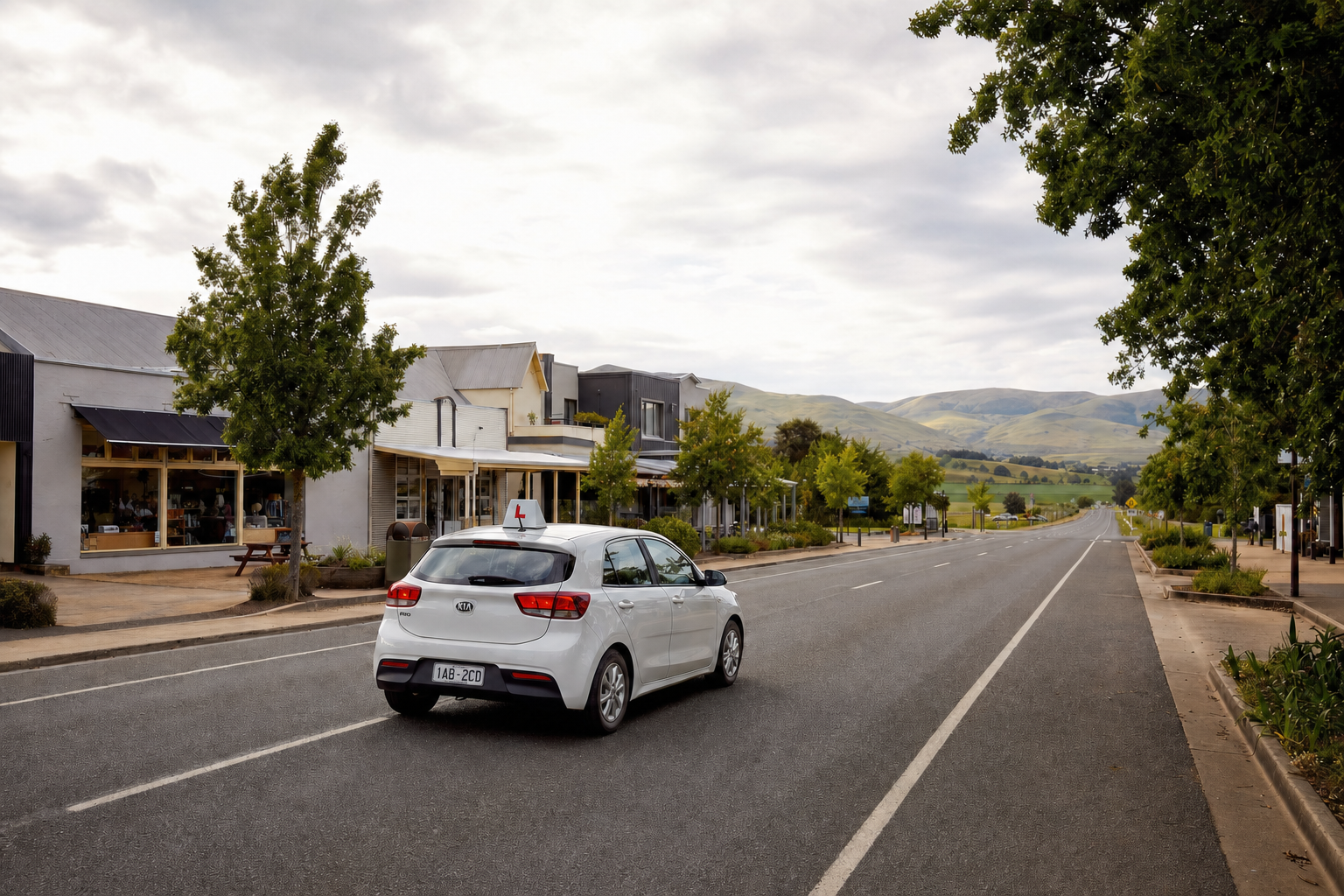 A white car with a learner sign drives down a quiet street lined with shops and trees, leading to distant green hills.