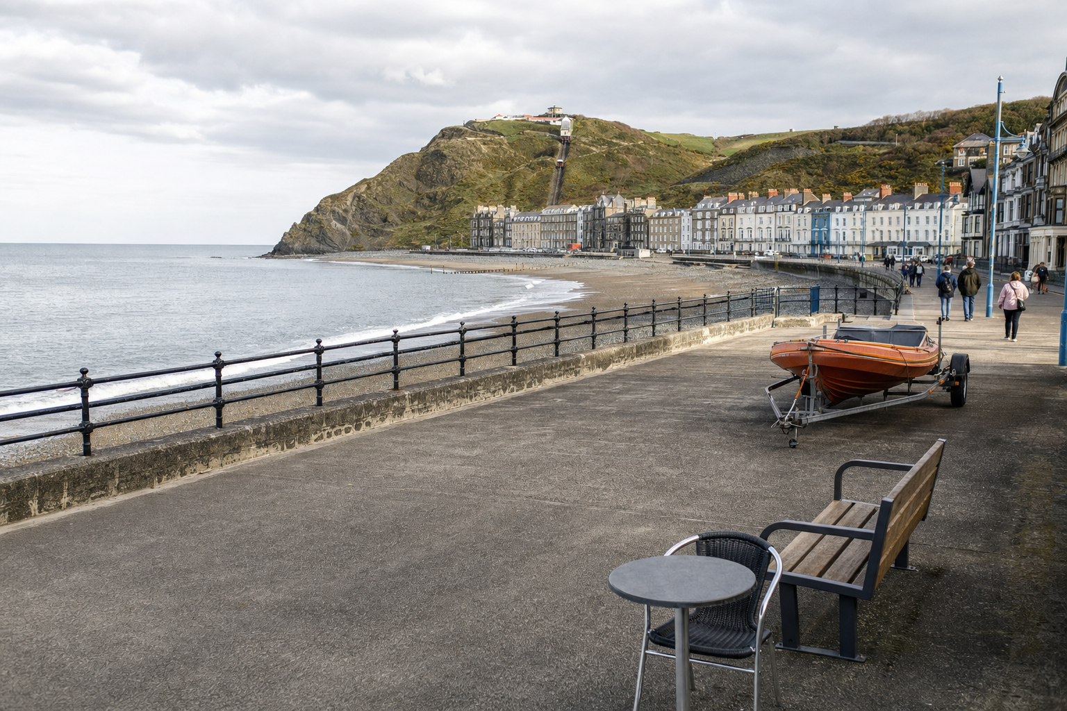 Coastal promenade with a small orange boat on a trailer, overlooking a beach and a row of houses in the background.